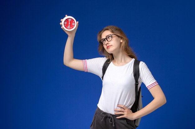 Front view of female student in white shirt wearing backpack and holding clocks on blue desk