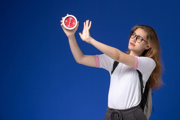 Front view of female student in white shirt wearing backpack and holding clocks on blue wall