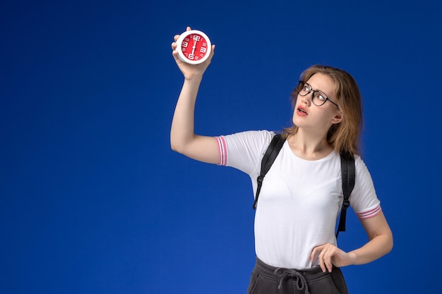 Front view of female student in white shirt wearing backpack and holding clocks on the blue wall