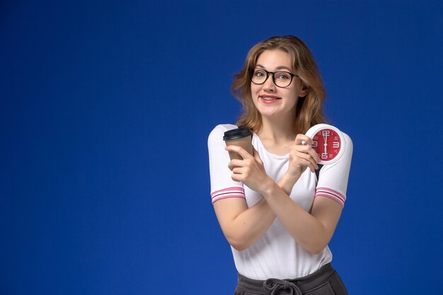 Front view of female student in white shirt wearing backpack and holding clocks and coffee on blue wall