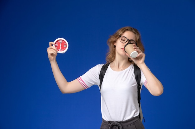 Front view of female student in white shirt wearing backpack and holding clocks and coffee on blue wall