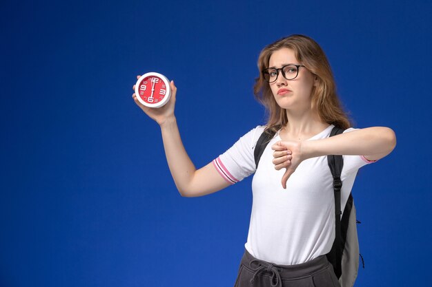 Front view of female student in white shirt wearing backpack and holding clocks showing unlike sign on the blue wall