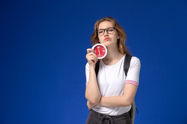 Front view of female student in white shirt wearing backpack and holding clocks with displeased expression on blue wall