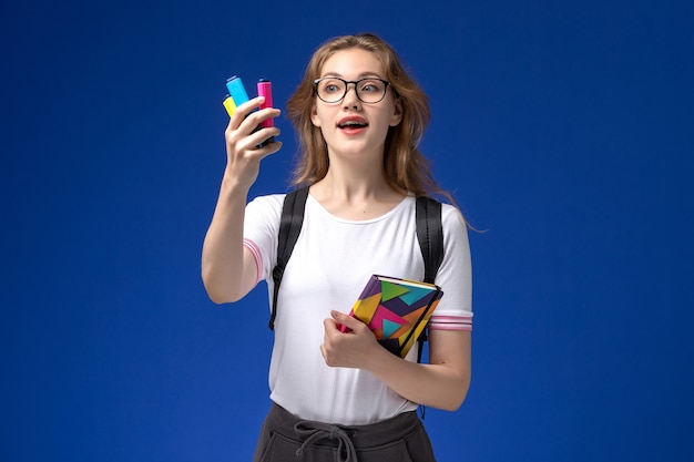 Front view of female student in white shirt wearing backpack and holding copybook with felt pens on the blue wall