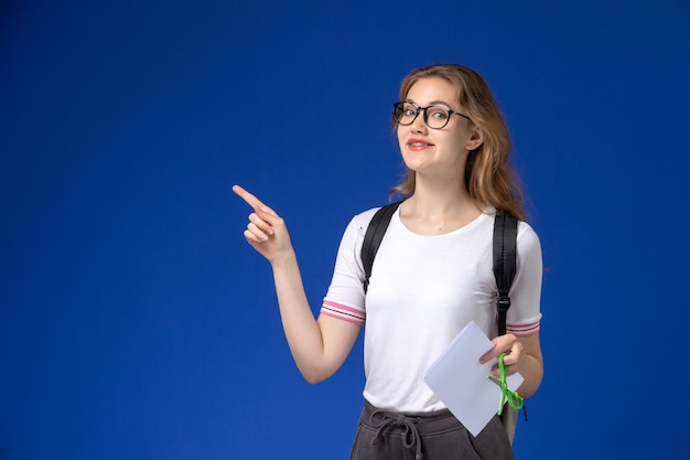 배낭을 착용하고 파란색 벽에 웃는 종이와 가위를 들고 흰 셔츠에 여성 학생의 전면보기
