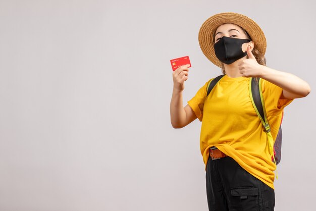 Front view of female traveller with backpack holding up card giving thumbs up on grey wall