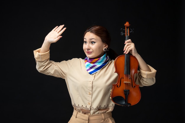 暗い壁に手を振っている彼女のバイオリンを保持している正面図の女性バイオリニストコンサートメロディー楽器女性パフォーマンス音楽感情プレイ