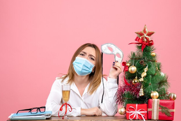 Front view of female worker in mask sitting behind her table with xmas presents on pink