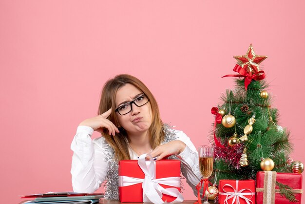 Front view of female worker sitting around christmas presents and tree on pink