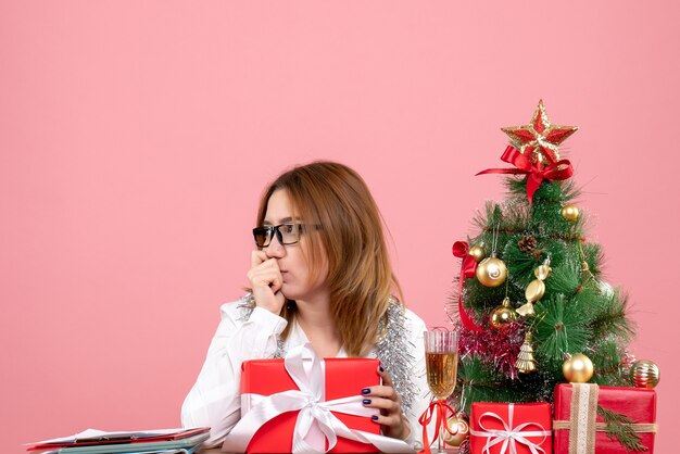 Front view of female worker sitting around christmas presents and tree on pink