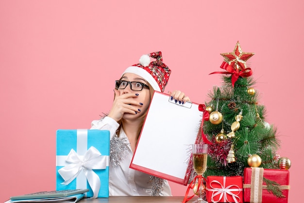 Front view of female worker sitting and holding file note around presents with shocked face on pink