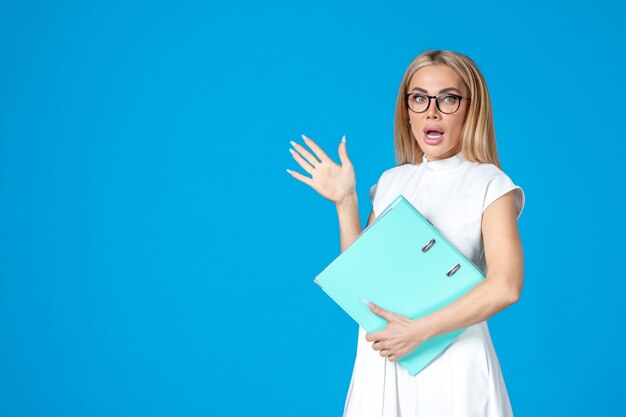 Front view of female worker in white dress holding blue folder on blue wall