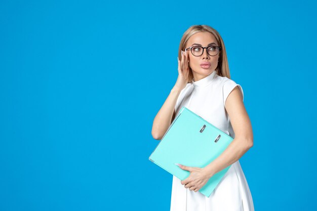 Front view of female worker in white dress holding blue folder on blue wall