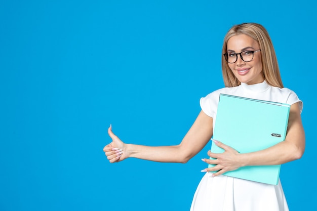 Front view of female worker in white dress holding blue folder on blue wall