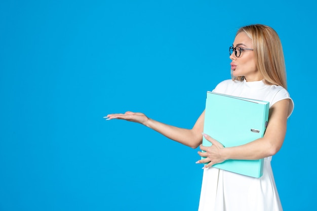 Front view of female worker in white dress holding blue folder on blue wall