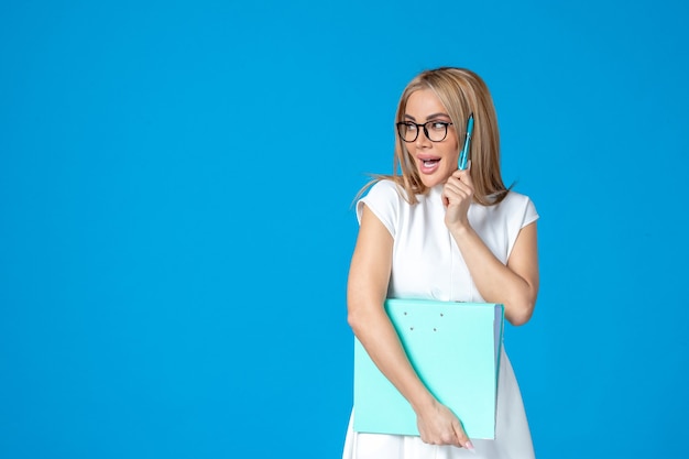 Front view of female worker in white dress holding blue folder on blue wall