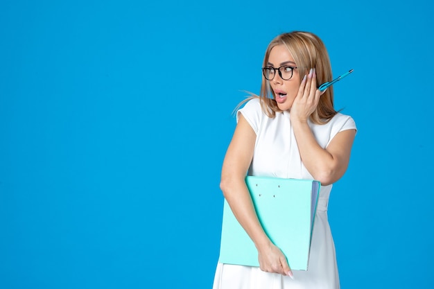 Front view of female worker in white dress holding blue folder on blue wall