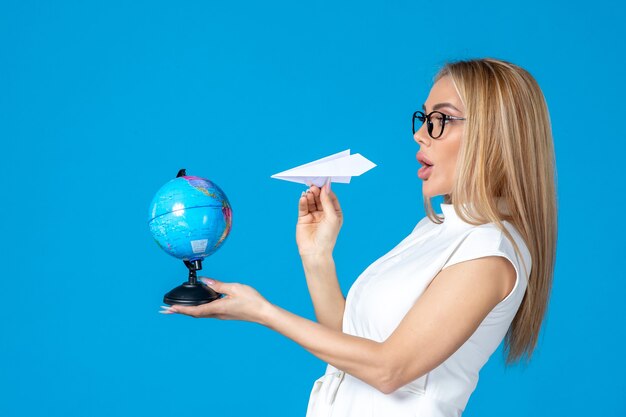 Front view of female worker in white dress holding earth globe and paper plane on blue wall