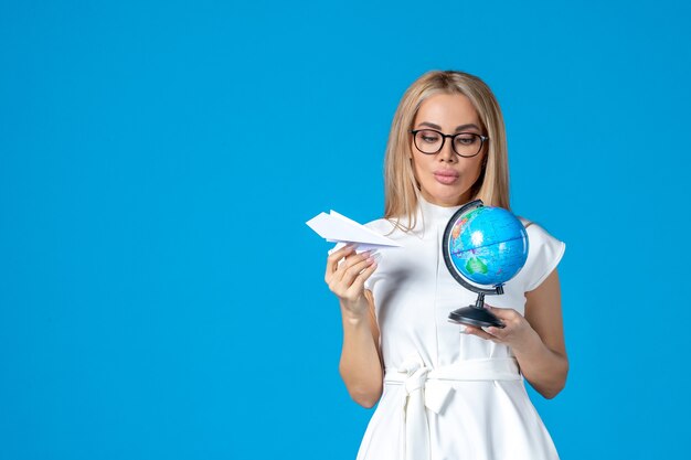 Front view of female worker in white dress holding earth globe and paper plane on blue wall