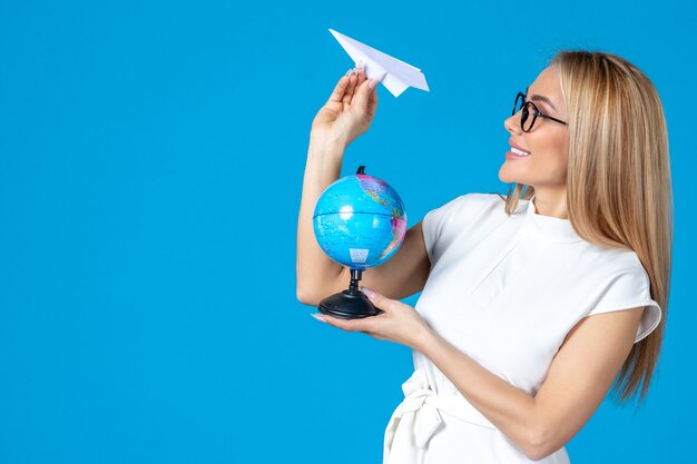 Front view of female worker in white dress holding earth globe and paper plane on blue wall