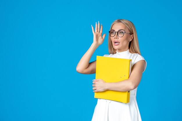 Front view of female worker in white dress holding folder and greeting someone on blue wall