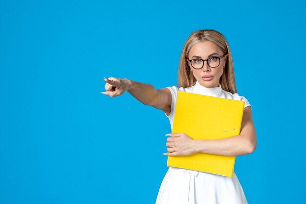 Front view of female worker in white dress holding folder and pointing on blue wall