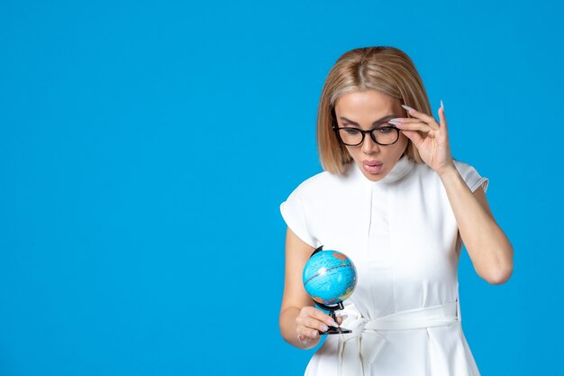 Front view of female worker in white dress holding little earth globe on blue wall