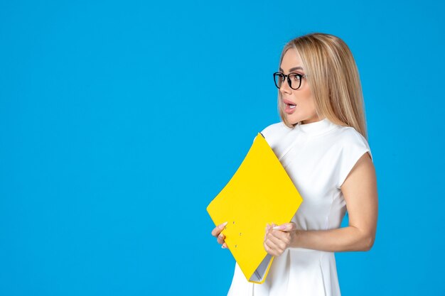 Front view of female worker in white dress holding yellow folder on blue wall