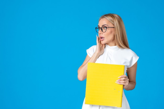 Front view of female worker in white dress holding yellow folder on blue wall