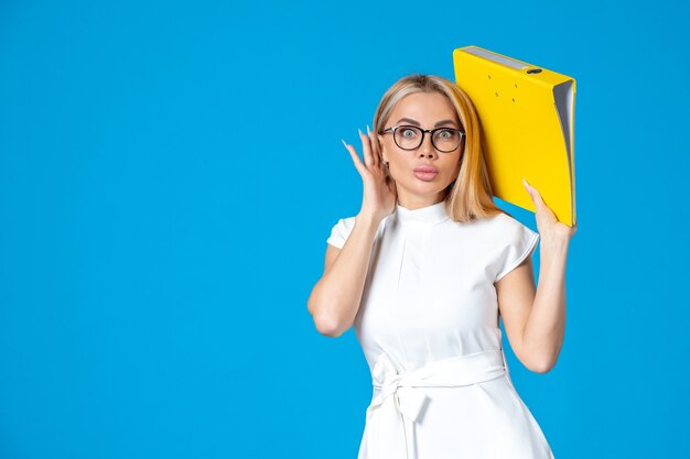 Front view of female worker in white dress holding yellow folder on blue wall