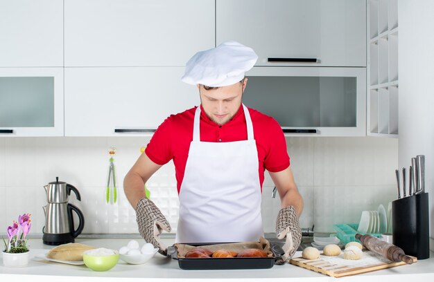 白いキッチンで焼きたてのペストリーを保持しているホルダーを身に着けた集中した男性シェフの正面図