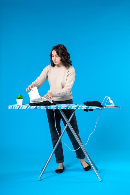 Front view of focused young woman standing behind the ironing board and holding cloth on blue wave surface