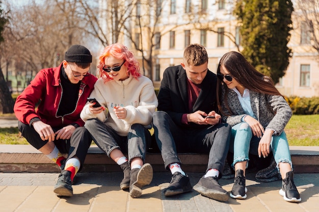 Front view of four friends together outdoors checking their smartphones