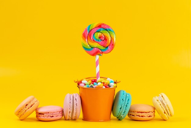 A front view french macarons alogn with lollipops and colorful candies on yellow, sweet color rainbow goody