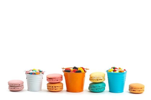 A front view french macarons along with colorful candies and marmalades inside colorful baskets on white, candy rainbow color