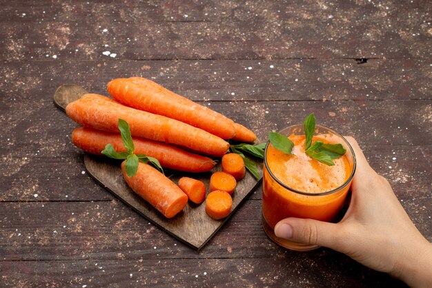 Front view fresh carrot juice inside long glass with leaf and along with fresh carrots on the brown desk 