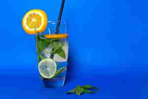 Free photo a front view fresh cold coktail with ice-cubes leaves citruses and black straw inside glass on blue