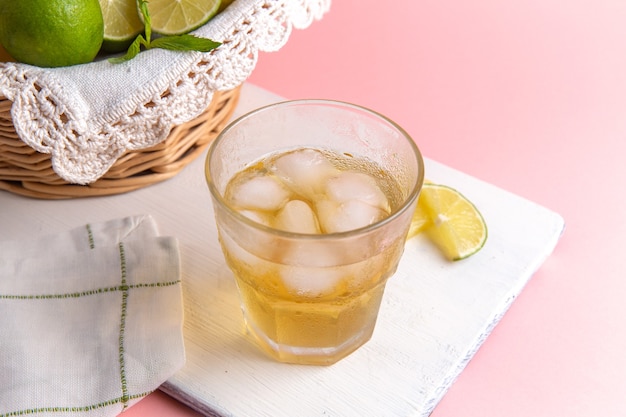 Front view of fresh cold lemonade with ice inside glass along with fresh lemons on pink desk