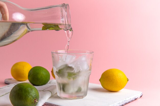 Front view of fresh cold lemonade with ice inside glass along with fresh lemons on pink wall