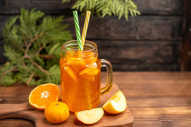 Вид спереди свежего фруктового сока в стакане с трубочками, яблоком и апельсином на деревянной разделочной доске