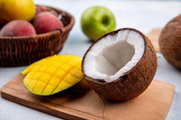 Front view of fresh fruits like sliced mango and half coconut on a wooden kitchen board with apples and peaches on a bucket on white surface