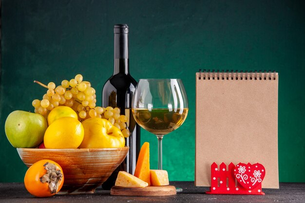 正面図木製ボウルリンゴの新鮮な果物マルメロレモンブドウ柿ワインボトルと緑のテーブルの木製ボードメモ帳にガラスチーズ