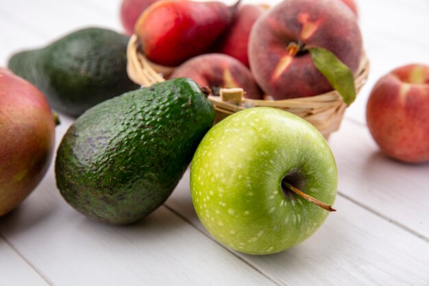 Front view of fresh peaches on a bucket with avocado and apple on a white surface