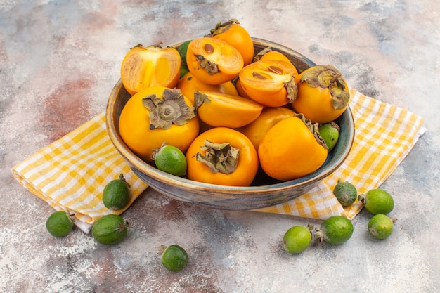 Front view fresh persimmons in a bowl yellow kitchen towel feijoa on nude background