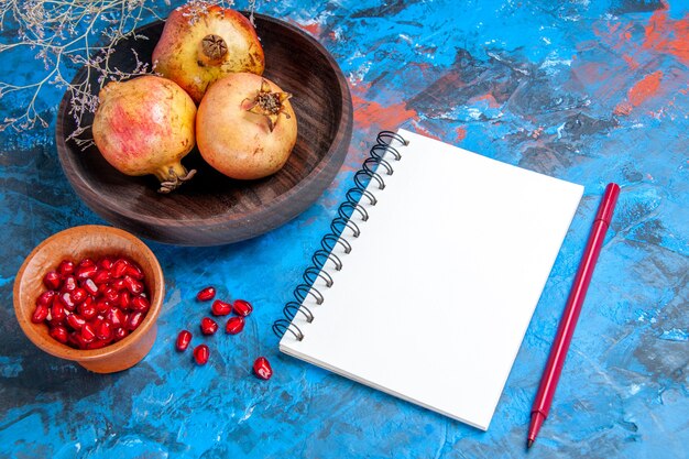 Front view fresh pomegranates in wooden bowl a bowl with pomegranate seeds a notebook red pen on blue