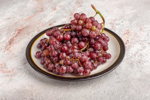 Vista frontale frutta fresca succosa e pastosa dell'uva rossa all'interno del piatto sulla superficie bianca