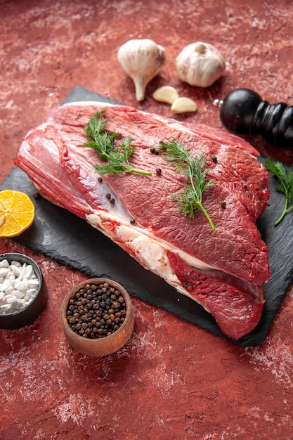 Front view of fresh red meat with green and pepper on black board knife garlics lemon spices wooden hammer lemon on oil pastel red background