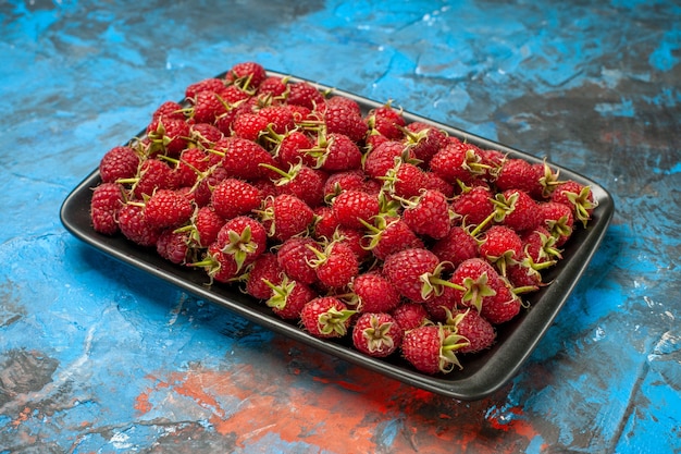 Вид спереди свежей красной малины внутри черного подноса на синем фоне цветных ягод диких спелых фруктов