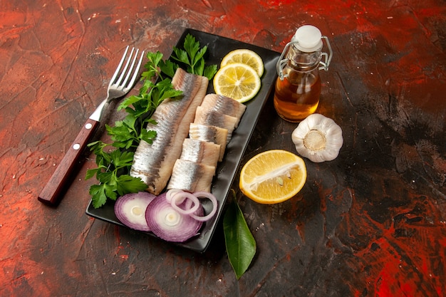 Вид спереди свежей нарезанной рыбы с зеленью, лимоном и луком внутри черной сковороды на темной фотографии закуски цвета морепродуктов