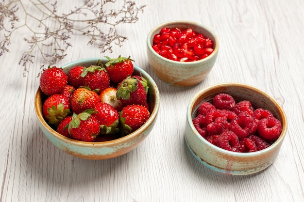 Вид спереди свежей клубники с малиной и гранатом на белом столе, ягоды, свежие фрукты, спелые спелые дикие